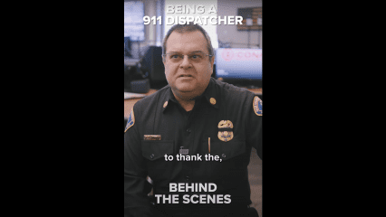 A uniformed fire department official is seated indoors, speaking on camera in what appears to be a dispatch or communications center. He is wearing a black uniform with a badge and nameplate, and monitors are visible in the background. Bold white text on the image reads: “BEING A 911 DISPATCHER” at the top, and “to thank the,” and “BEHIND THE SCENES” near the bottom, indicating a tribute to 911 dispatchers.